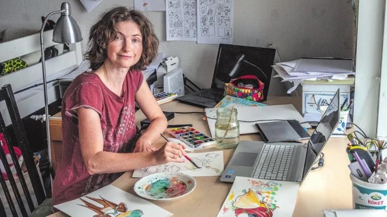 Tanja vid sitt arbetsbord med sina redskap – storyboard på väggen, akvarellfärger, ritplatta och dator.