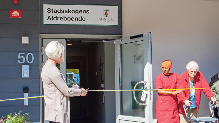 På bild syns bland andra undersköterskan Benny Bendén i blå klädsel samt enhetschefen Lina Sjögren uppe till höger. Till vänster i bild ses nämndens ordförande Eva-Lotta Pamp (M) i samtal med en boende.